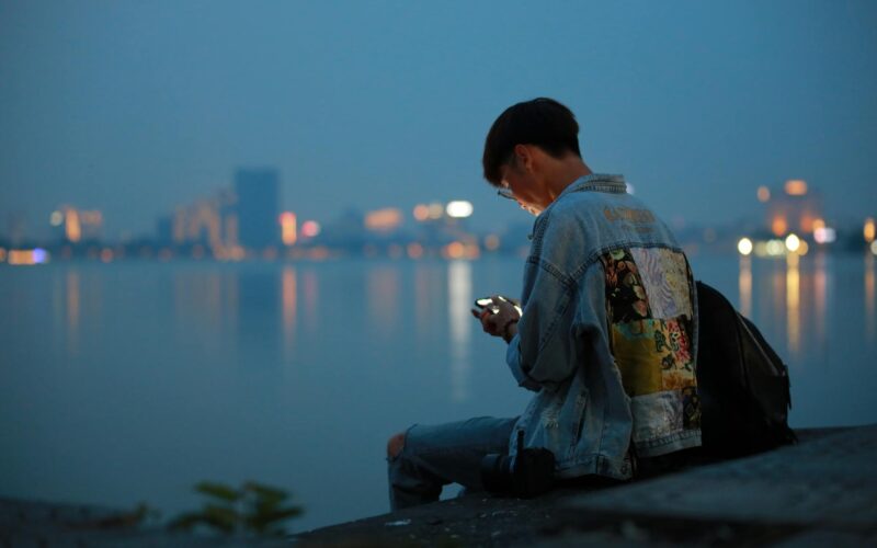 teen in denim jacket using phone by waterfront